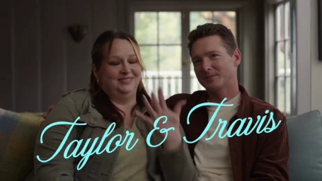 A smiling woman and man sitting together on a couch, with the woman holding up her hand showing a ring.