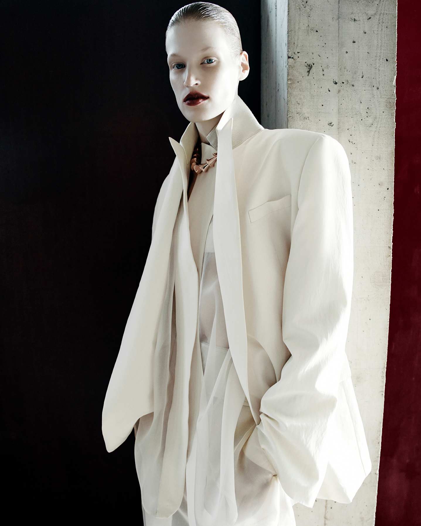 Portrait of a fair-skinned woman in a light beige suit and sheer top, posing in front of a dark wall and a textured light-colored wall.