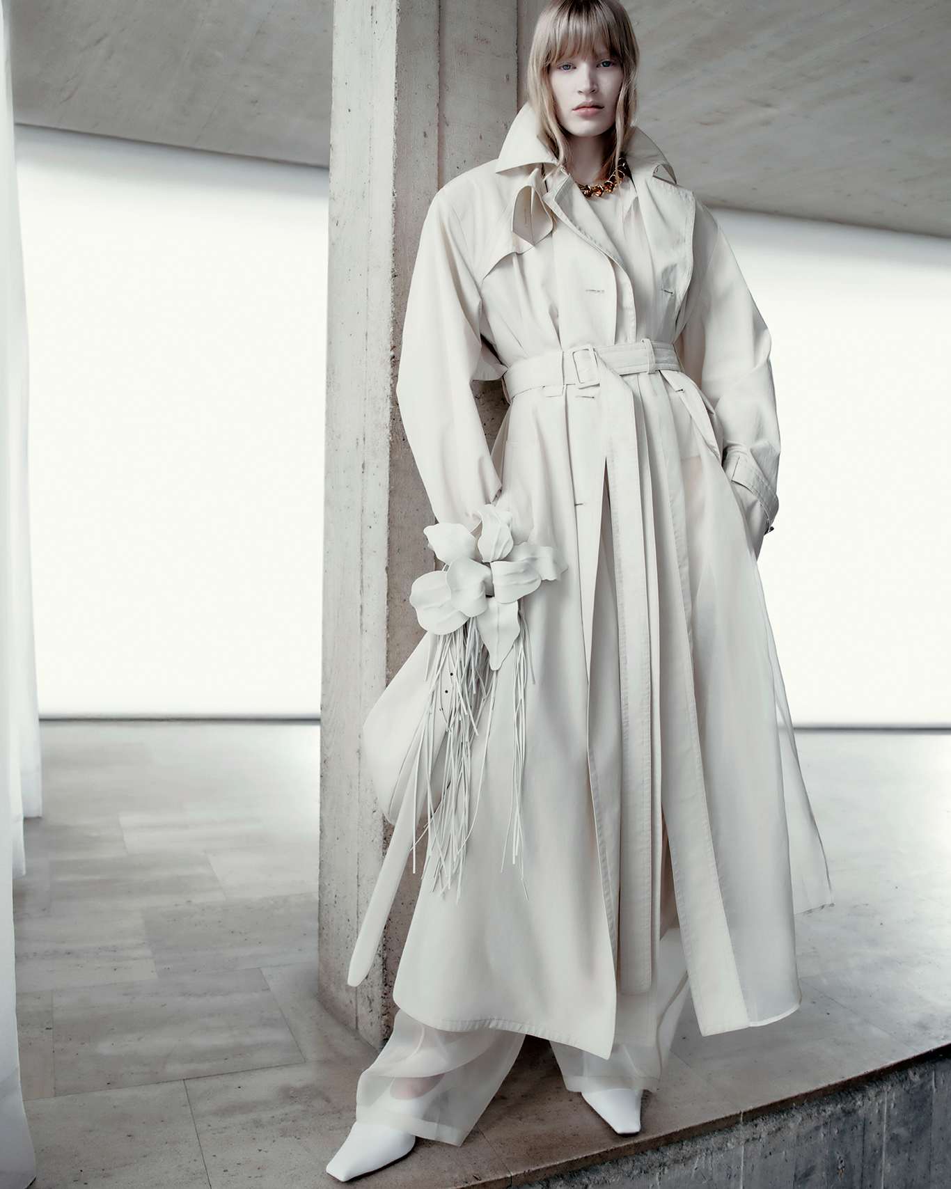 Indoor fashion shot featuring a blonde woman wearing a white trench coat, wide-leg pants, white boots, and holding a bouquet of flowers.