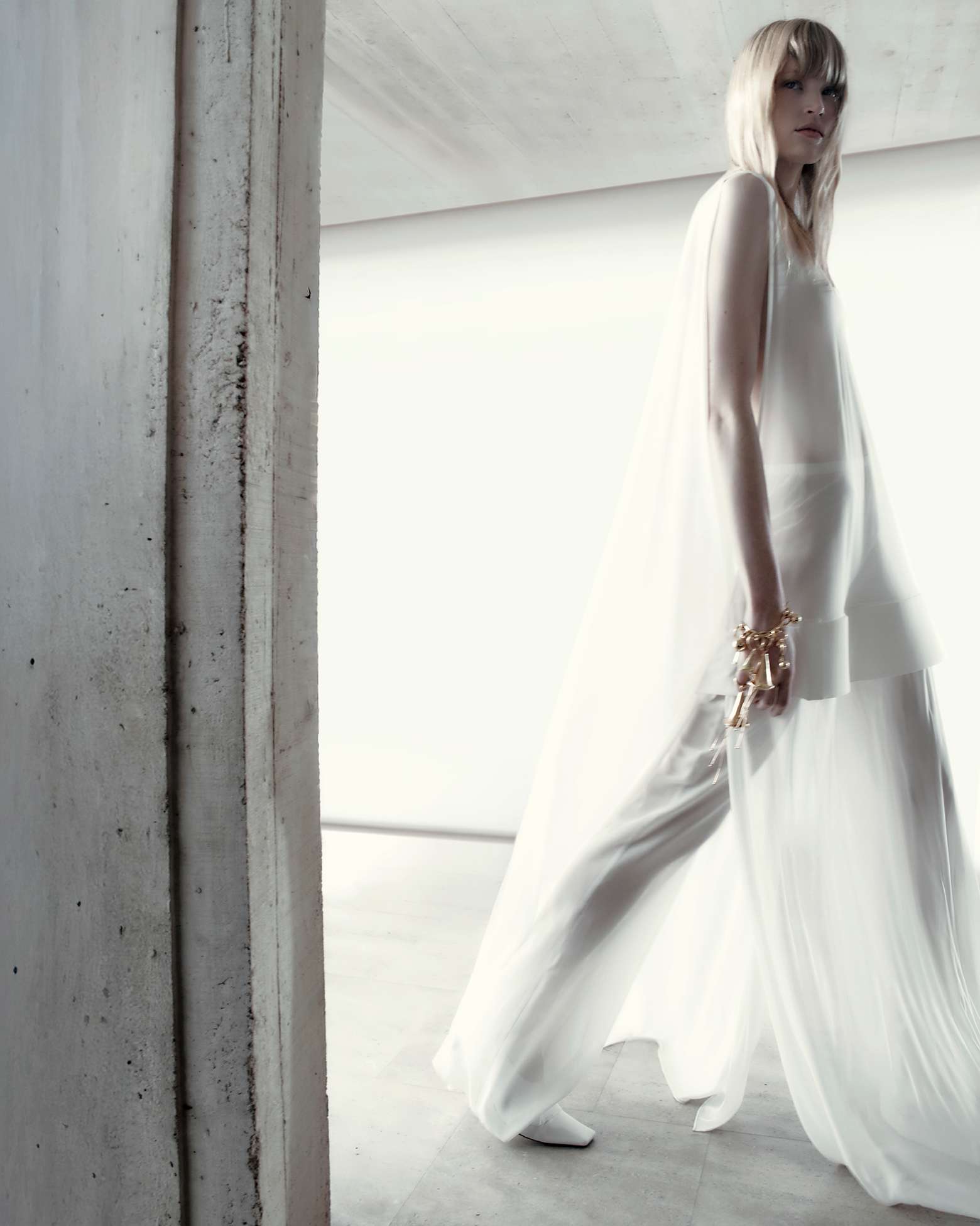 Fashion shot of an adult woman with blonde hair wearing a white outfit, gold bracelets, and white heels in an indoor environment with concrete walls.