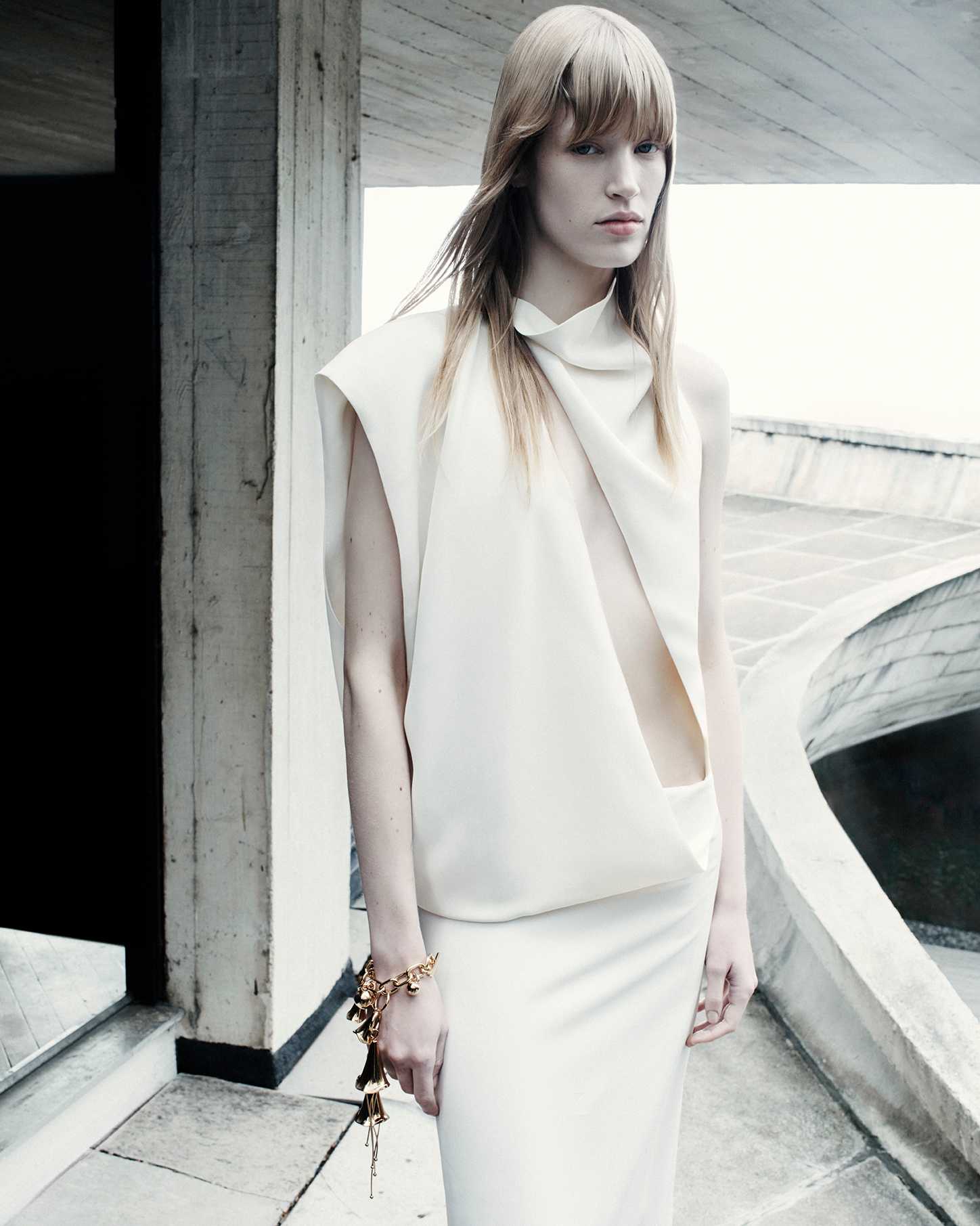 Model in white draped sleeveless dress wearing a gold charm bracelet standing against architectural concrete