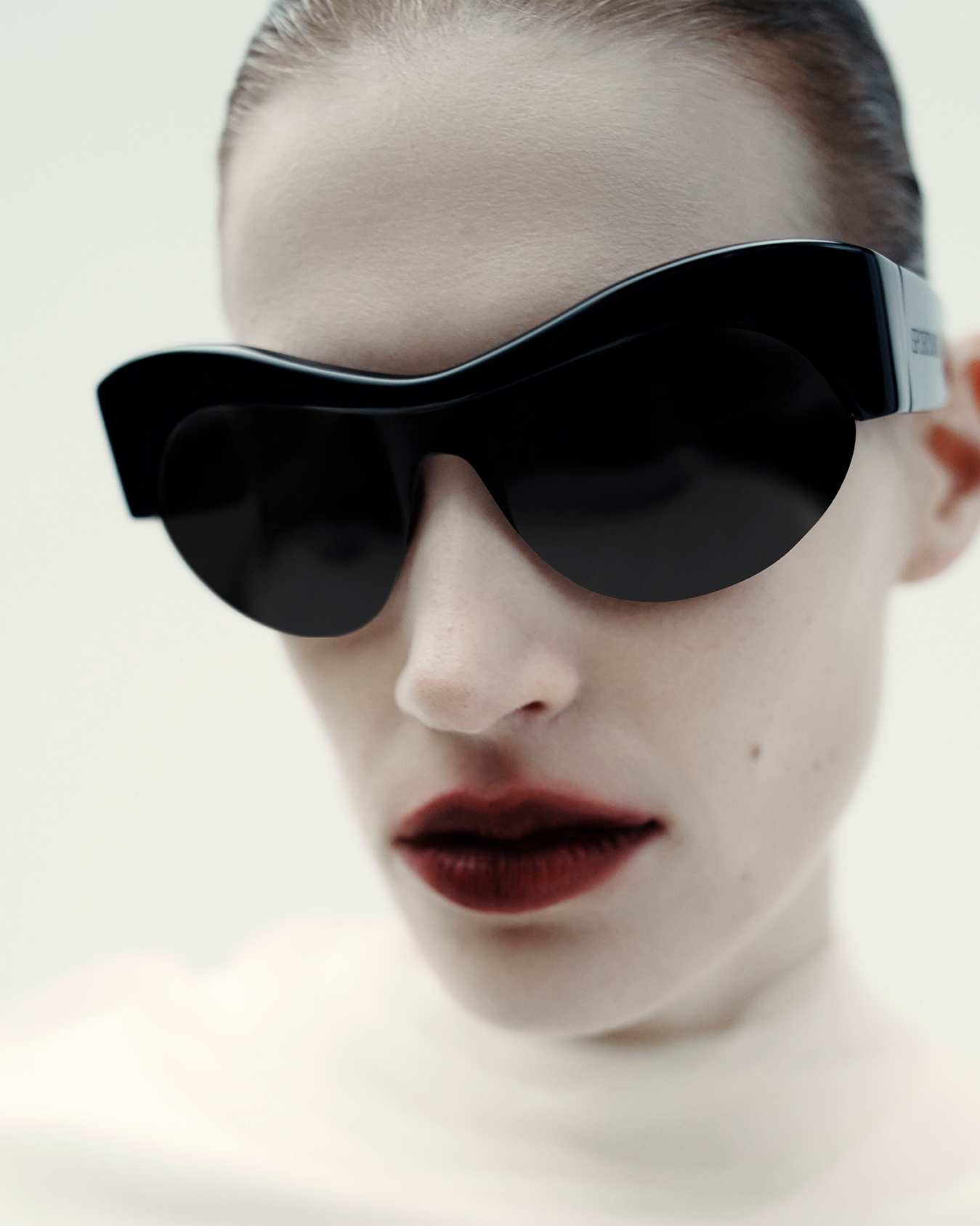 Close-up of a stylish adult female wearing black sunglasses and dark lipstick against a light background in a studio setting.