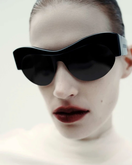 Close-up of a stylish adult female wearing black sunglasses and dark lipstick against a light background in a studio setting.