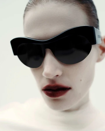 Close-up of a stylish adult female wearing black sunglasses and dark lipstick against a light background in a studio setting.