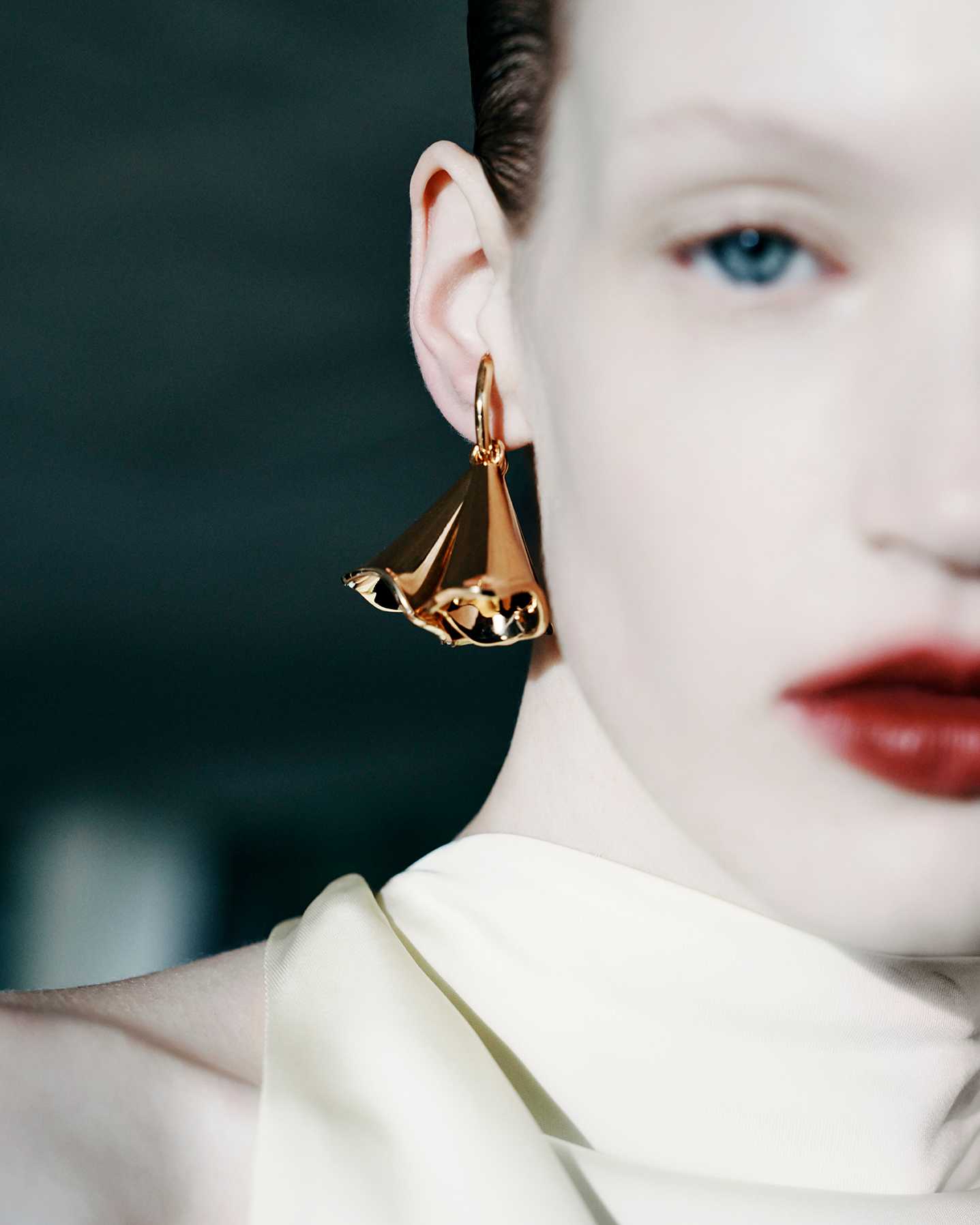 Close-up of a fair-skinned woman with red lipstick and a gold hoop earring with a sculptural pendant against a dark background.