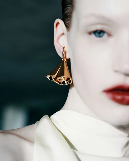 Close-up of a fair-skinned woman with red lipstick and a gold hoop earring with a sculptural pendant against a dark background.