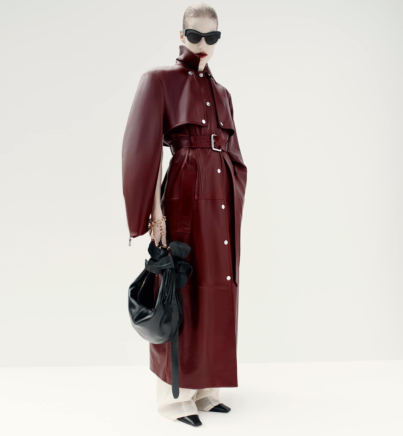 Fashionable woman in a burgundy coat and sunglasses stands against a white backdrop with a black handbag, exuding modern elegance.