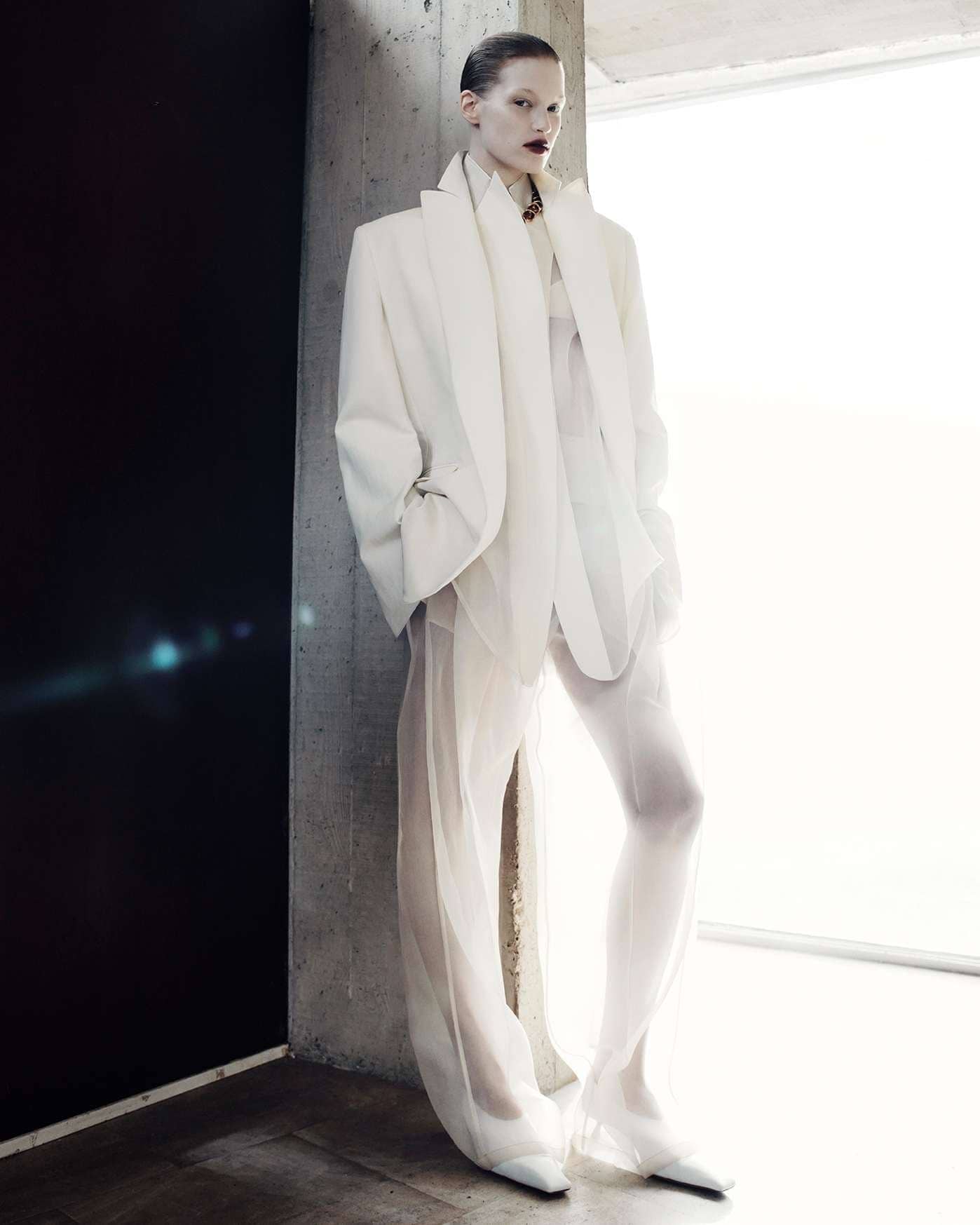 Fashion editorial shot of a slender woman in a white pantsuit and sheer pants standing in front of a grey concrete wall and window.