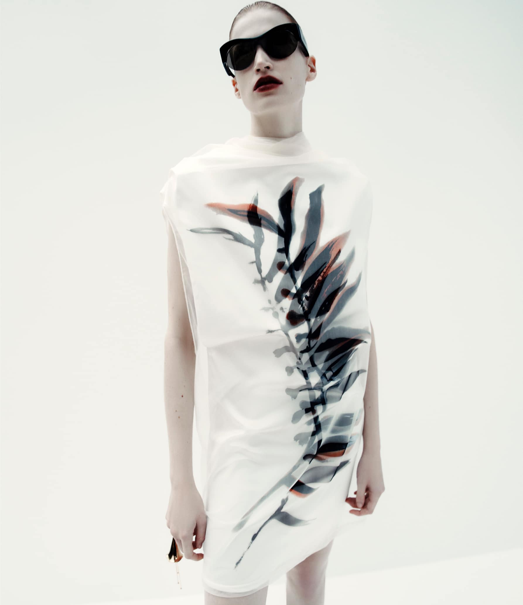Fashion portrait of a woman wearing a translucent dress with a leaf design and dark sunglasses against a bright white backdrop.