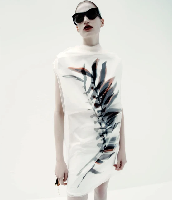 Fashion portrait of a woman wearing a translucent dress with a leaf design and dark sunglasses against a bright white backdrop.