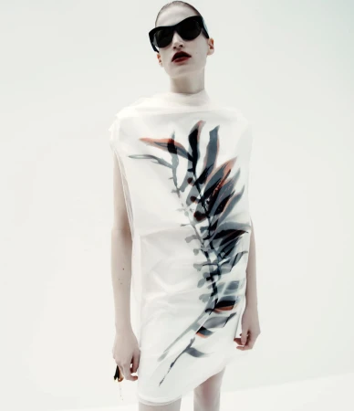 Fashion portrait of a woman wearing a translucent dress with a leaf design and dark sunglasses against a bright white backdrop.