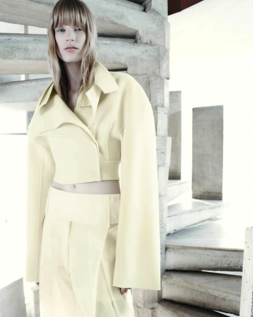 Blonde woman in pale yellow crop top and skirt poses in a bright interior with concrete spiral staircase, looking directly at the camera.