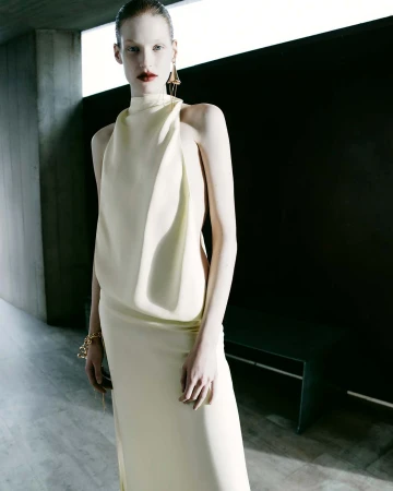 An adult female poses indoors wearing a high-necked sleeveless cream-colored dress with gold jewelry, under a concrete structure.