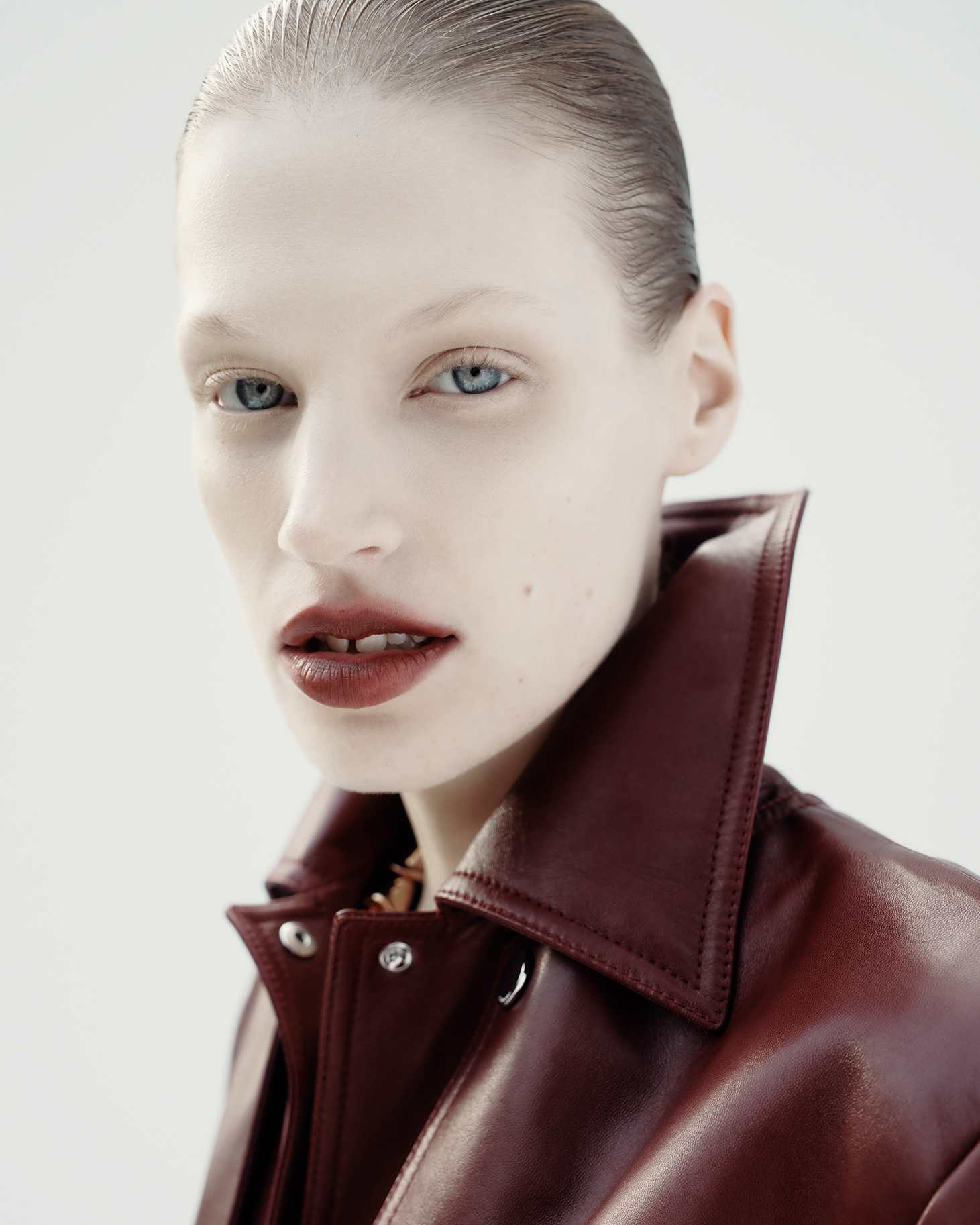 Close-up portrait of a fair-skinned adult woman with slicked-back hair, wearing a burgundy leather jacket against a bright background.