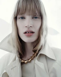 Close-up portrait of a blonde model wearing a cream-colored collared coat and a gold statement necklace against a bright white background.