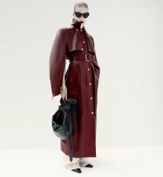 Model wearing a structured burgundy leather coat and sunglasses holding a black handbag against a white background