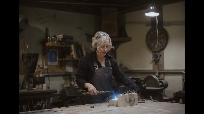 Adult female artisan in a workshop using a torch on metal, wearing safety glasses and an apron, with shop tools and equipment in view.