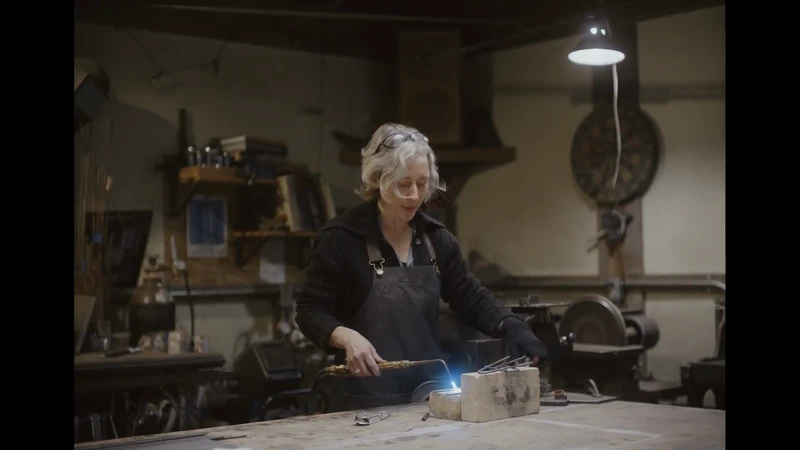 Adult female artisan in a workshop using a torch on metal, wearing safety glasses and an apron, with shop tools and equipment in view.