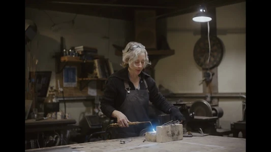An artisan woman wearing a dark apron welding metal in a workshop under a pendant light for KitchenAid branded content.