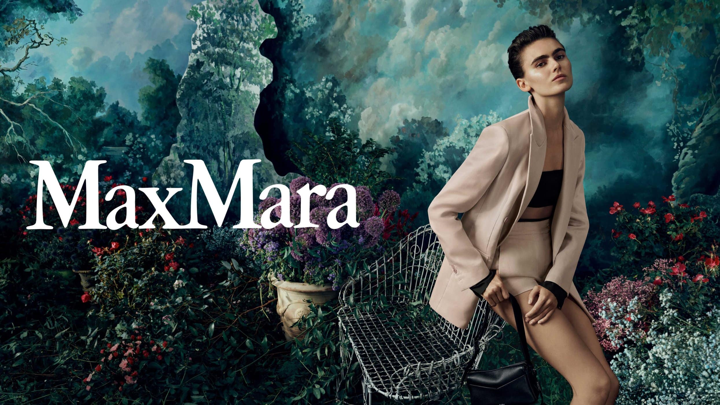 Fashion editorial featuring a woman in a beige blazer and shorts, seated in a wire chair against an outdoor garden backdrop, promoting MaxMara.