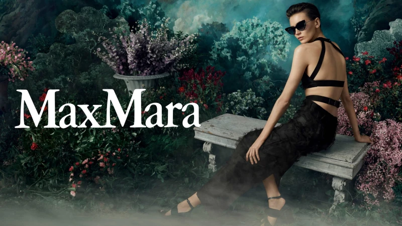 A woman in black fashion wear seated on a bench in a lush green garden with the Max Mara logo visible on the left side.