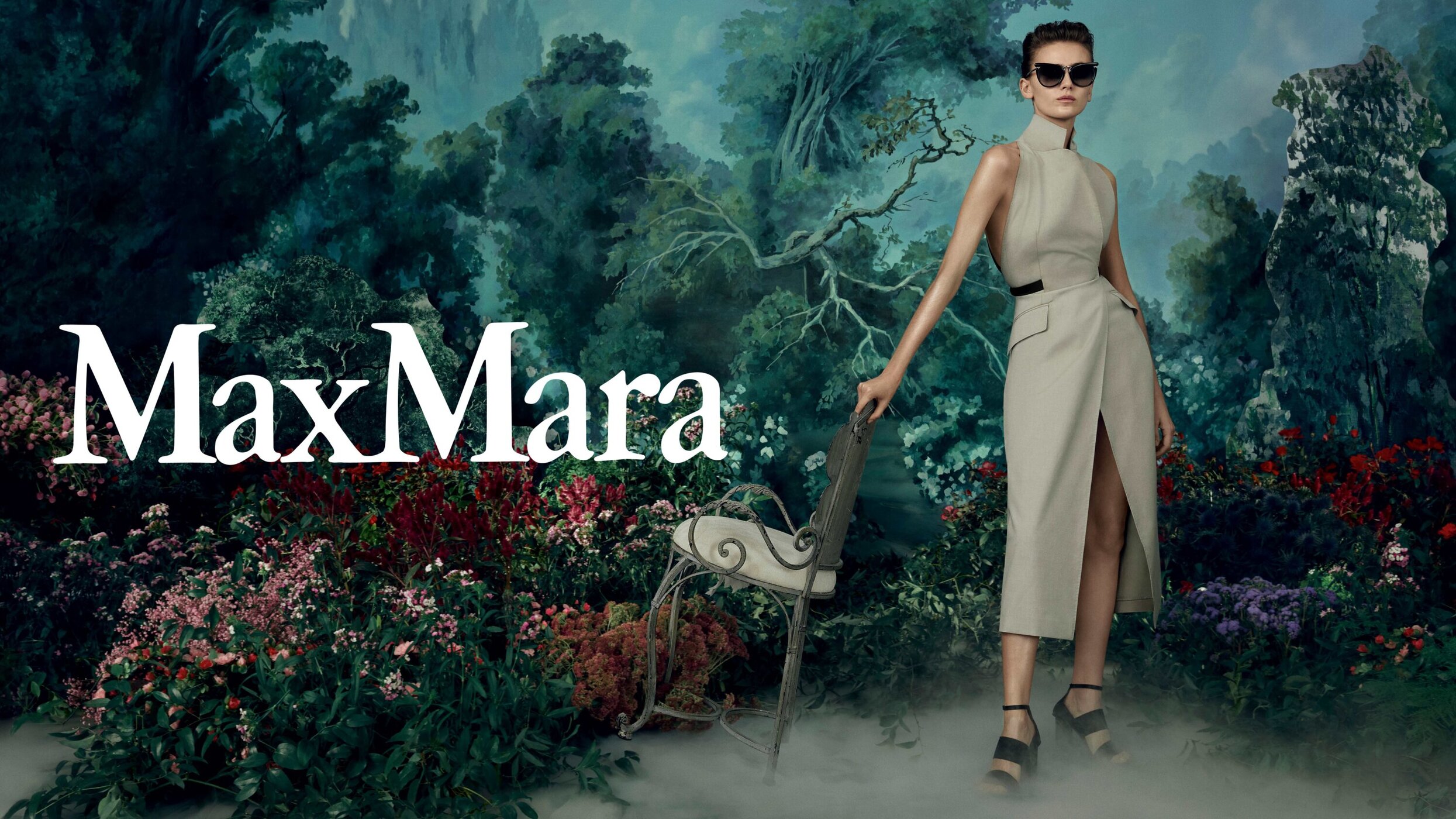 Fashion ad featuring woman in light beige dress and sunglasses, seated chair in a garden-like setting with floral foreground and backdrop.