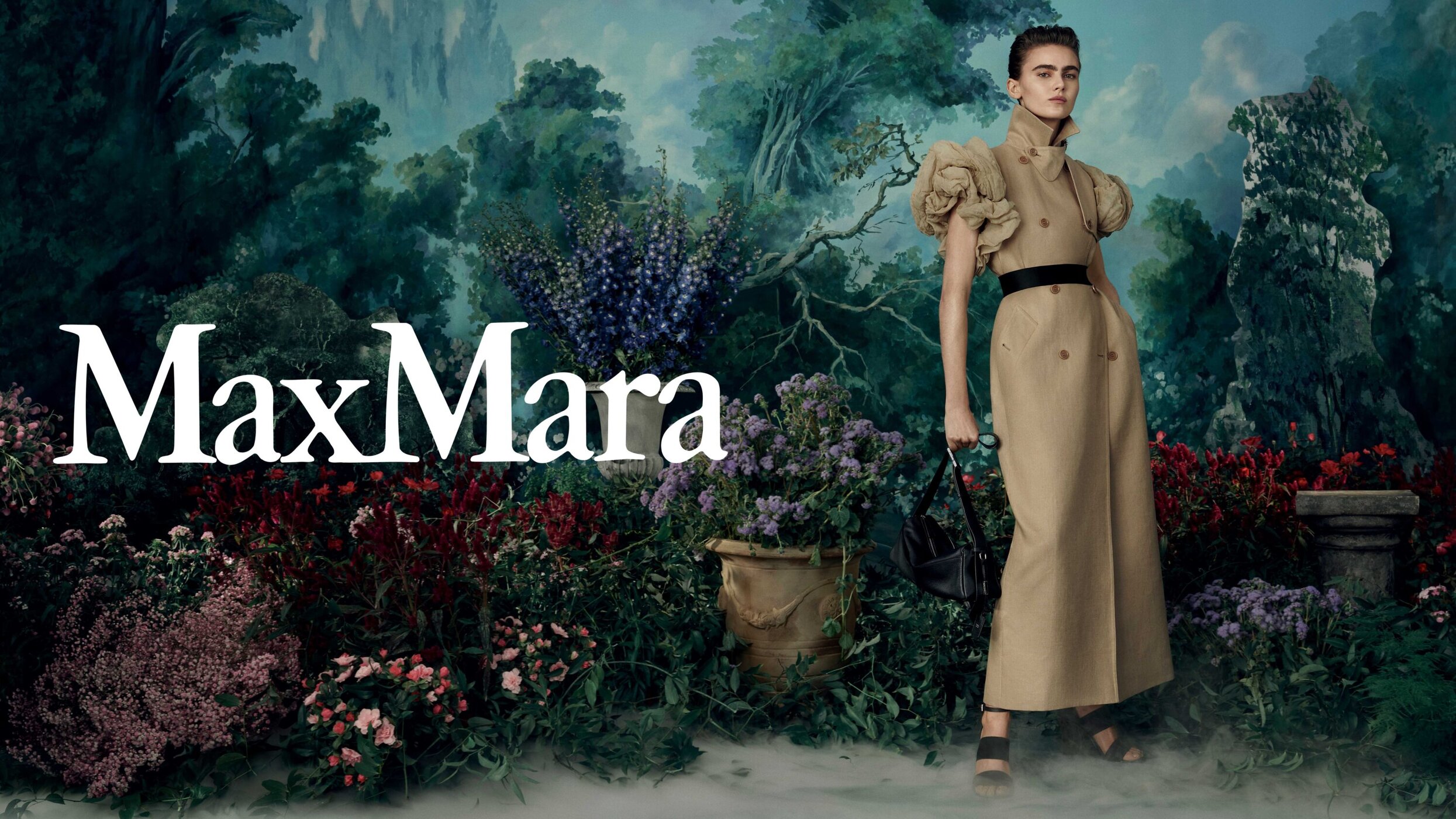 An adult woman models a Max Mara trench coat with voluminous sleeves in an outdoor setting with lush greenery and painterly backdrop.