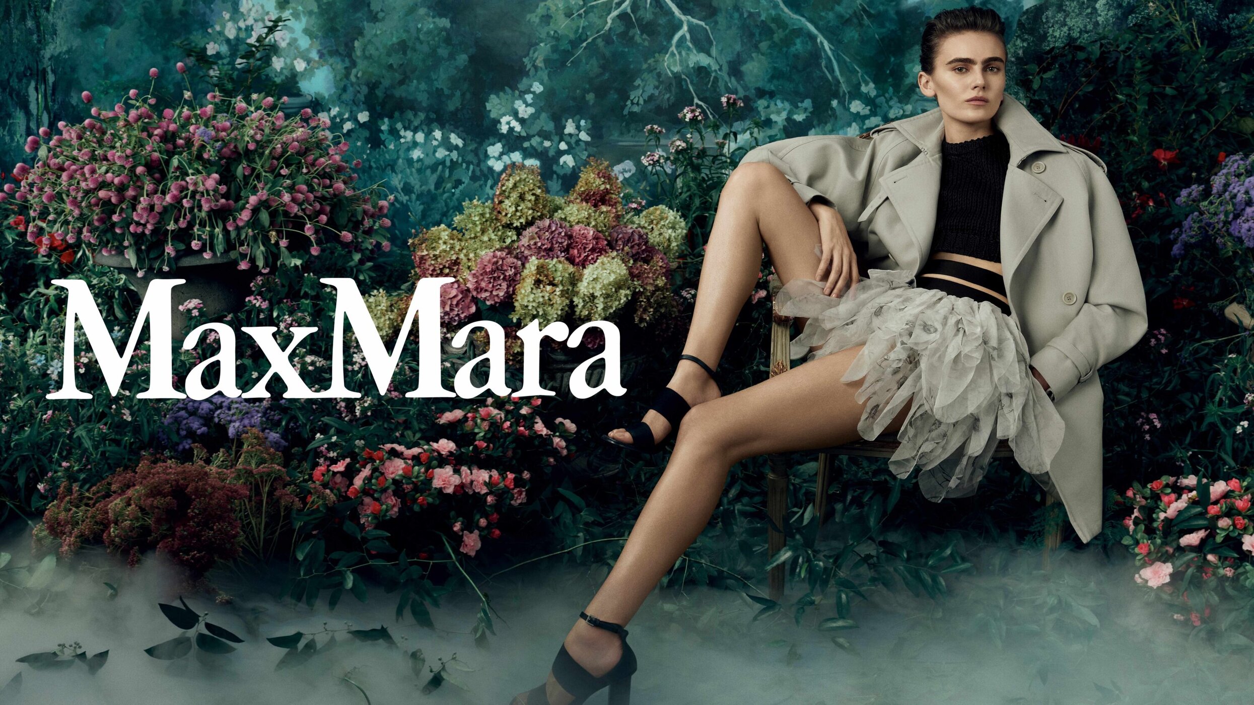 Model seated on a chair in a lush garden setting wearing a tan trench coat, black top, and tiered skirt for Max Mara.