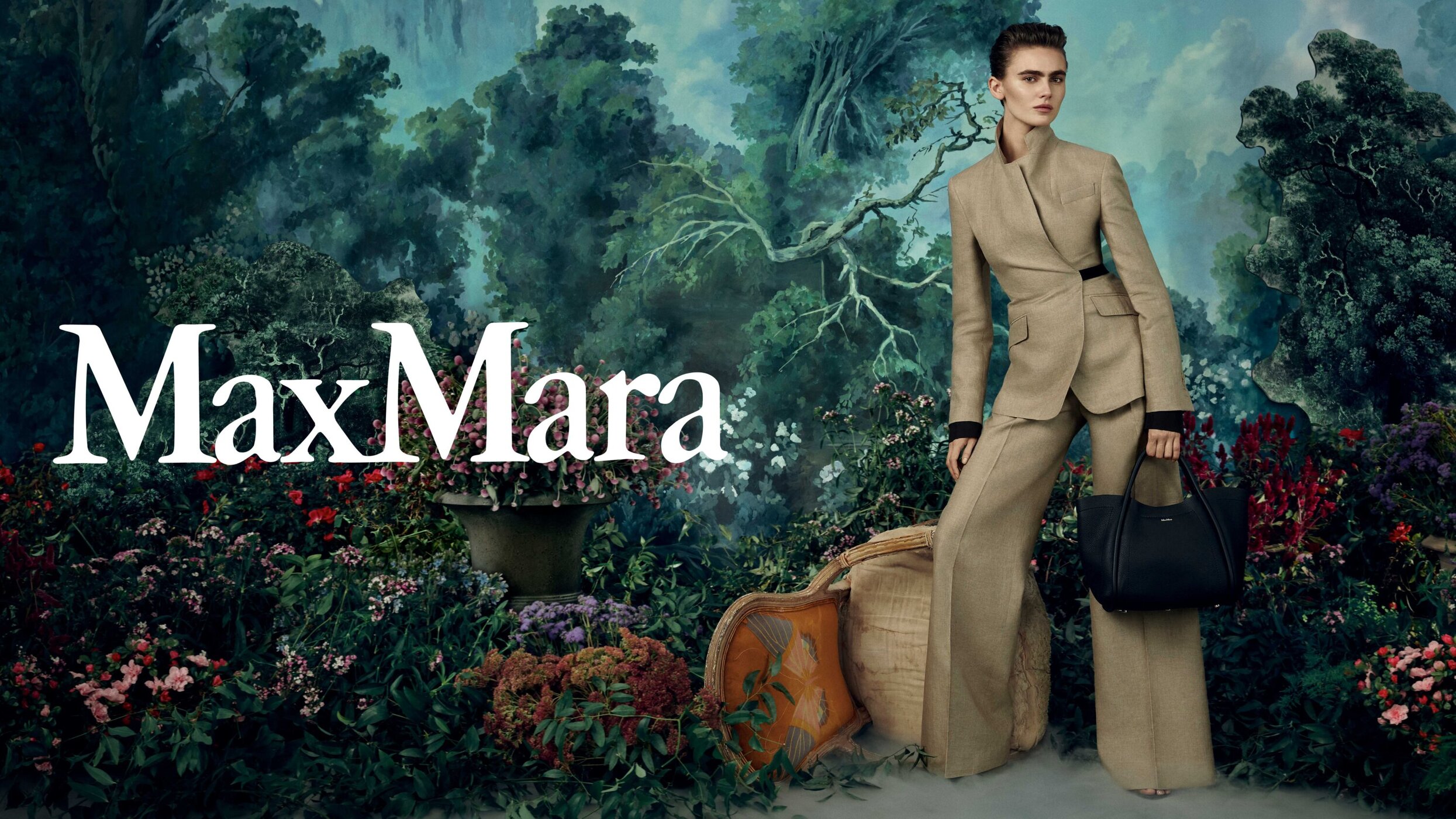 Elegant woman in a tan suit with a black handbag posing in front of a painterly garden backdrop for a Max Mara fashion campaign.