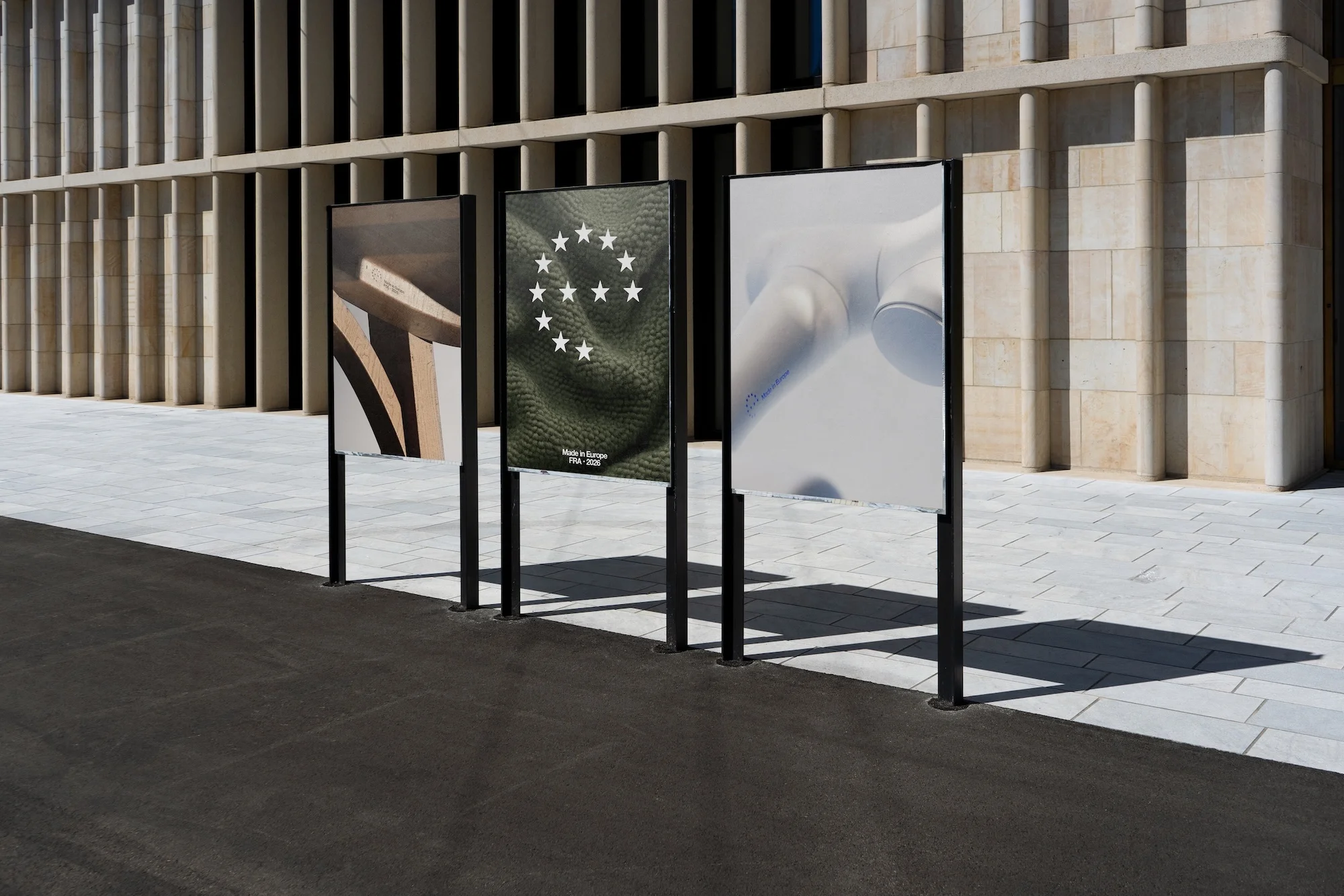 Three advertising posters on black metal frames stand outdoors against a modern building with stone and concrete columns during the day.