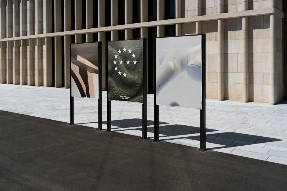 Three advertising posters on black metal frames stand outdoors against a modern building with stone and concrete columns during the day.