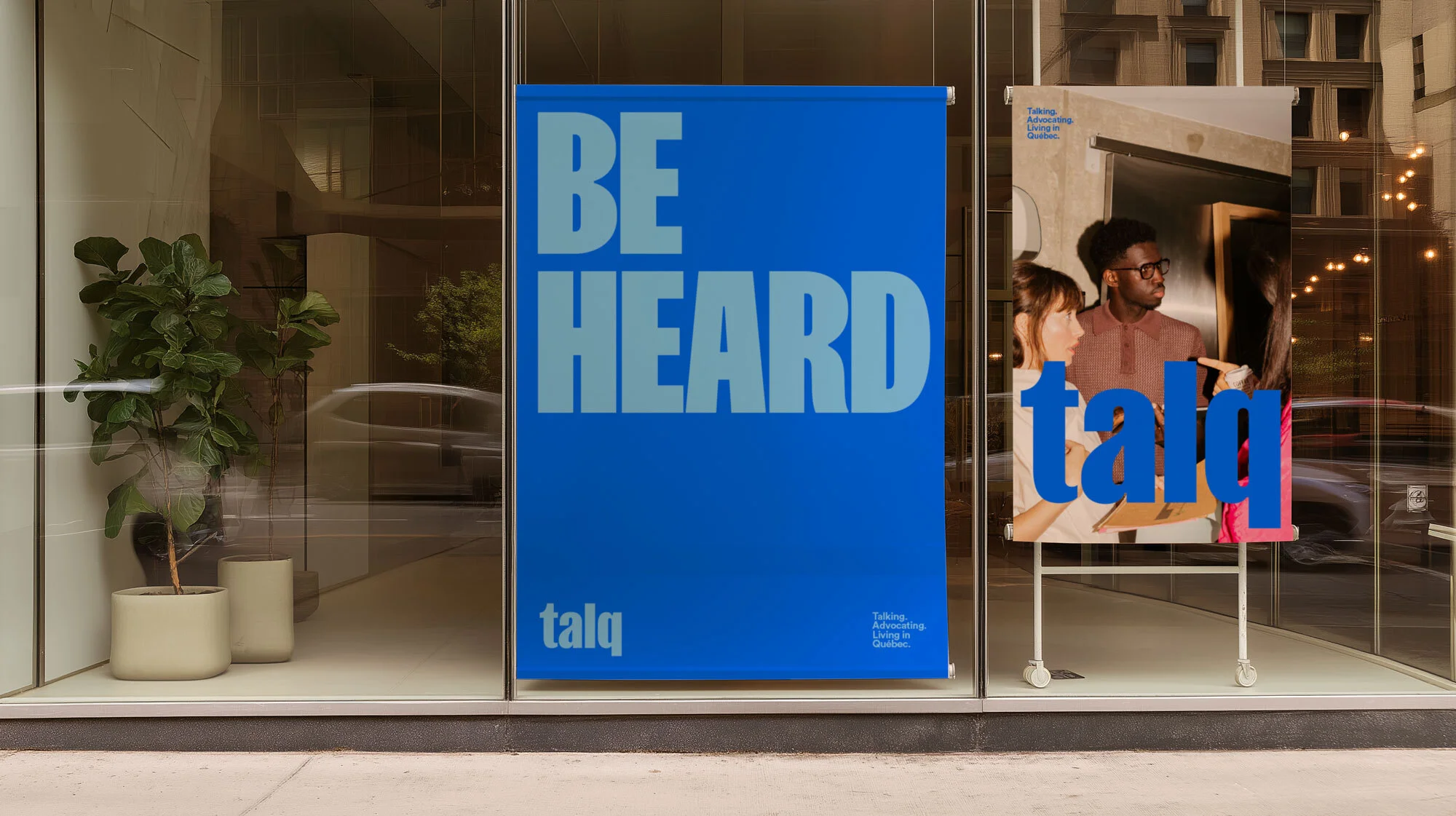 Eye-level outdoor shot of two marketing posters inside a storefront glass window with a plant in a pot in the window display.