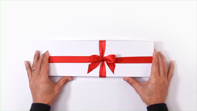 A person presents a long white gift box tied with a shiny red ribbon against a white background, with a focus on the box and hands.