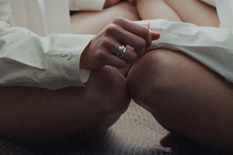 Close-up on two people's legs intertwined, wearing white shirts with an embroidered 'S', focus on a hand with simple metal rings.