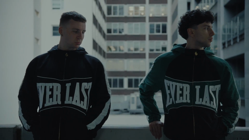 Two adult males stand outdoors wearing matching Everlast branded hooded jackets, set against an urban backdrop with tall buildings.