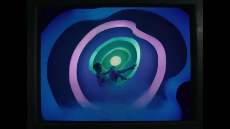 A person reclining inside a glowing, concentric tunnel installation with blue, purple, and green illumination.