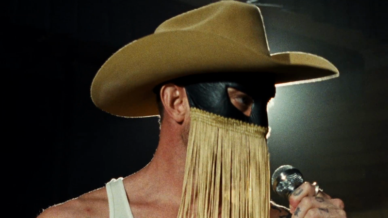 Close-up of an adult male wearing a cowboy hat and mask while singing into a microphone in a dark indoor environment.