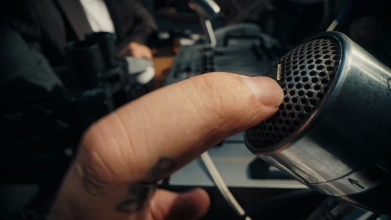 Close up shot of a man's hand turning on a vintage microphone. The man is wearing a suit coat, and there is a soundboard in background.