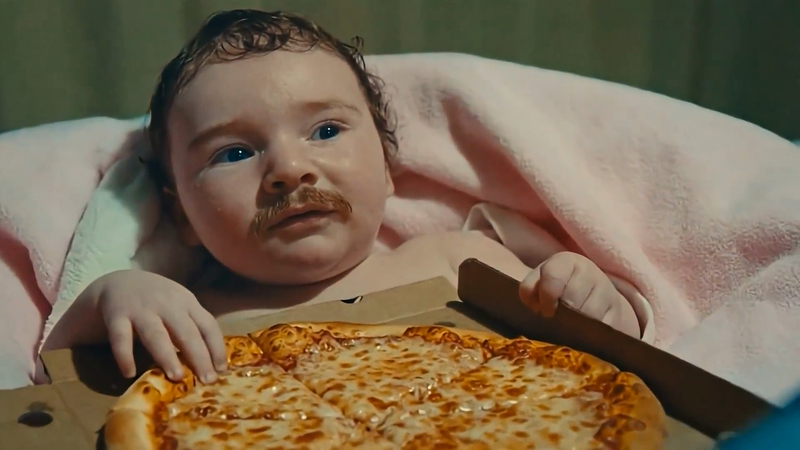 A baby with a drawn-on mustache lies under a pink blanket next to a pizza in a box.