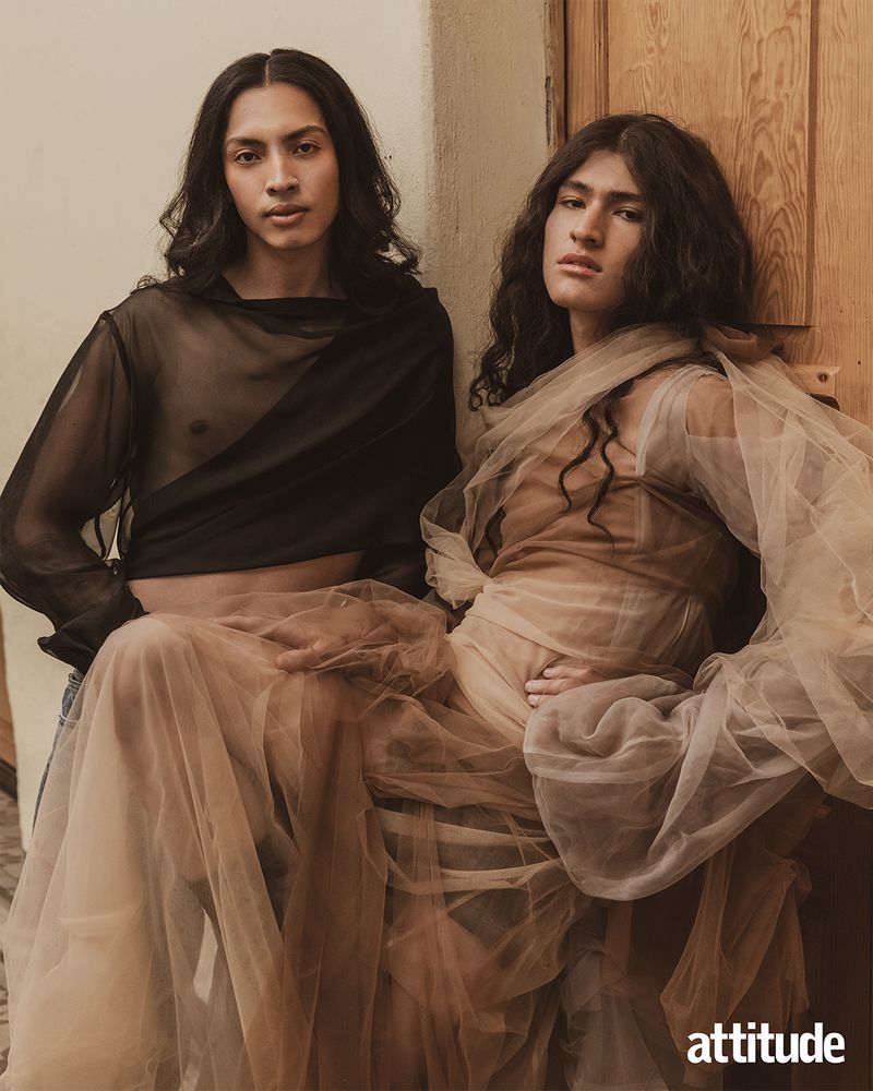 Two young adult males in sheer tops pose indoors, with one in a black top and the other in a beige draped top, next to a wooden door.