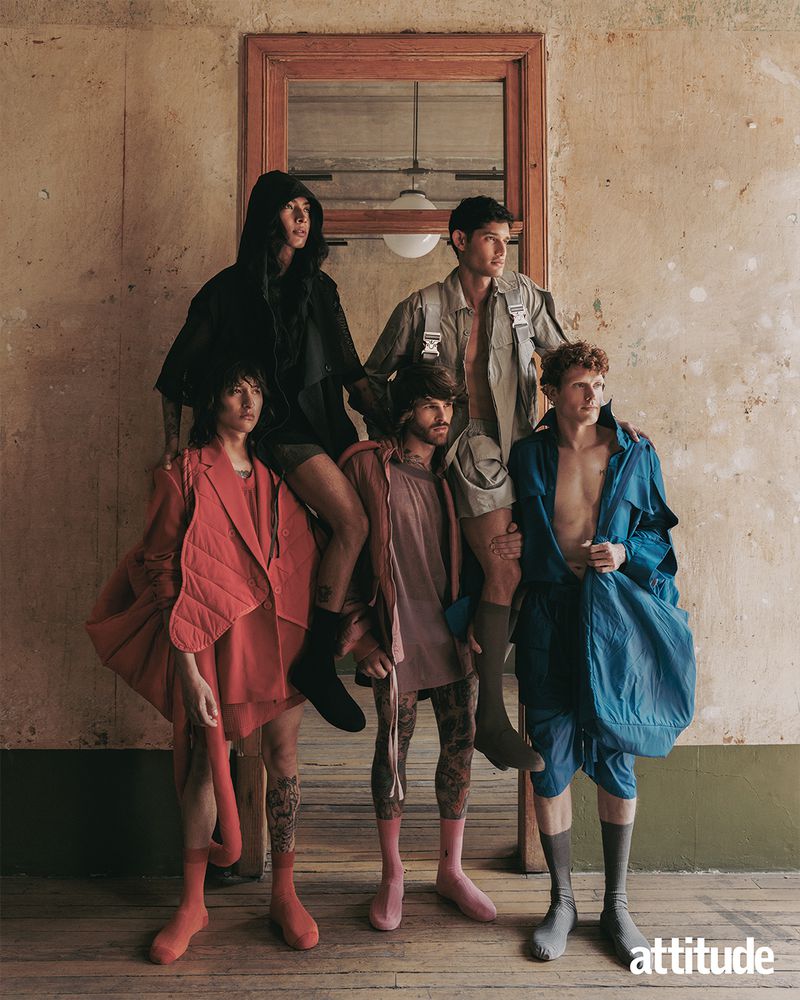 Editorial fashion shot of five young adult men in colorful outfits and knee socks posed indoors against a textured wall and window.