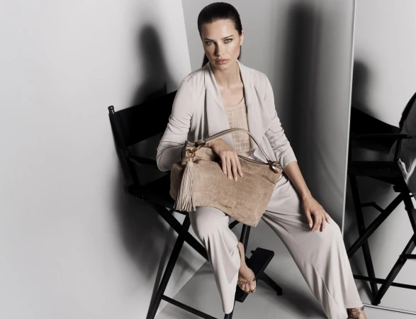 Adult woman poses in studio wearing a light tan-colored pants suit holding a handbag, seated on a director's chair.