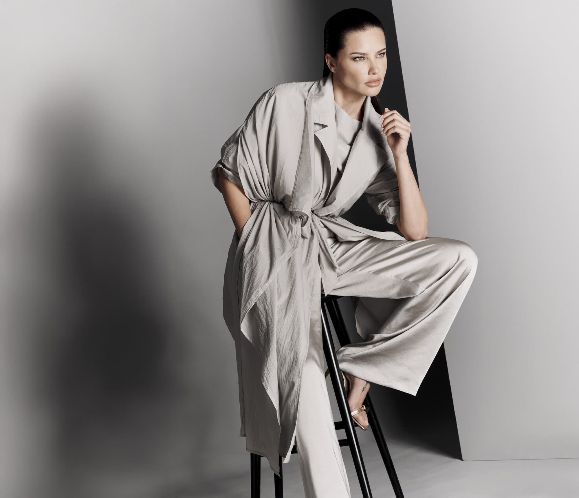 Elegant adult woman in a gray robe-like top and wide-leg trousers, seated on a black stool against a two-tone gray backdrop, fashion.