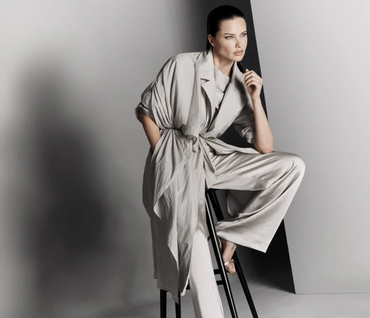Elegant adult woman in a gray robe-like top and wide-leg trousers, seated on a black stool against a two-tone gray backdrop, fashion.