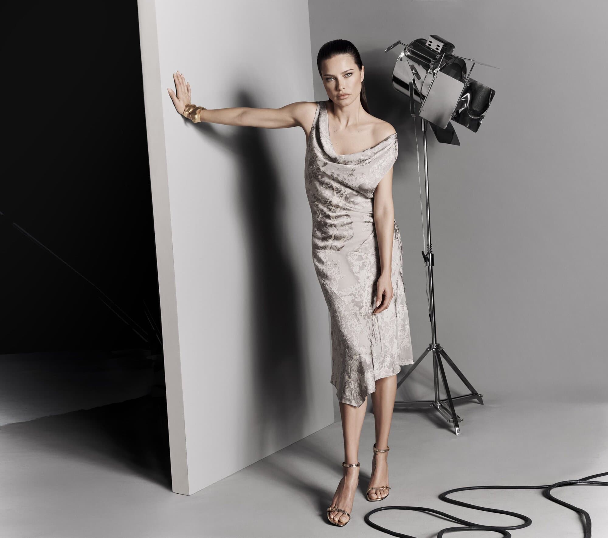 A full shot of an adult female fashion model in a dress posing in a photo studio with studio lighting and power cords on the floor.