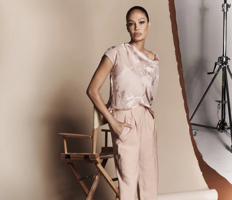 A woman poses in a studio wearing a light pink top and pants, standing near a director's chair, with a neutral color background.