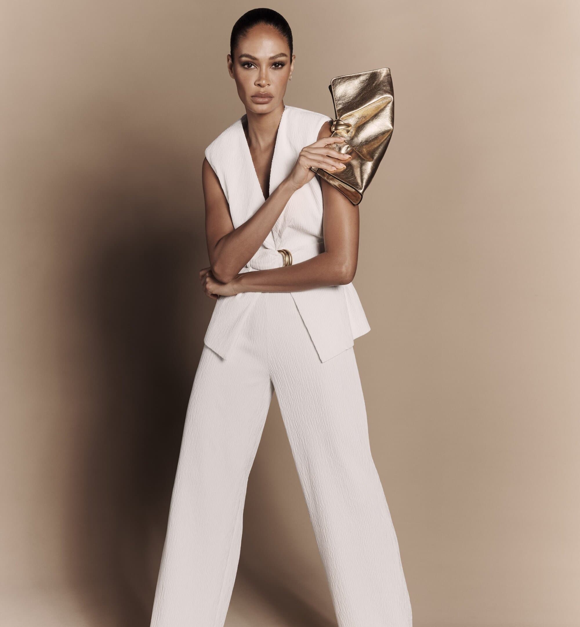 Elegant woman in a white sleeveless suit poses with a gold clutch in a studio setting. Fashionable, sleek look. Neutral color palette.