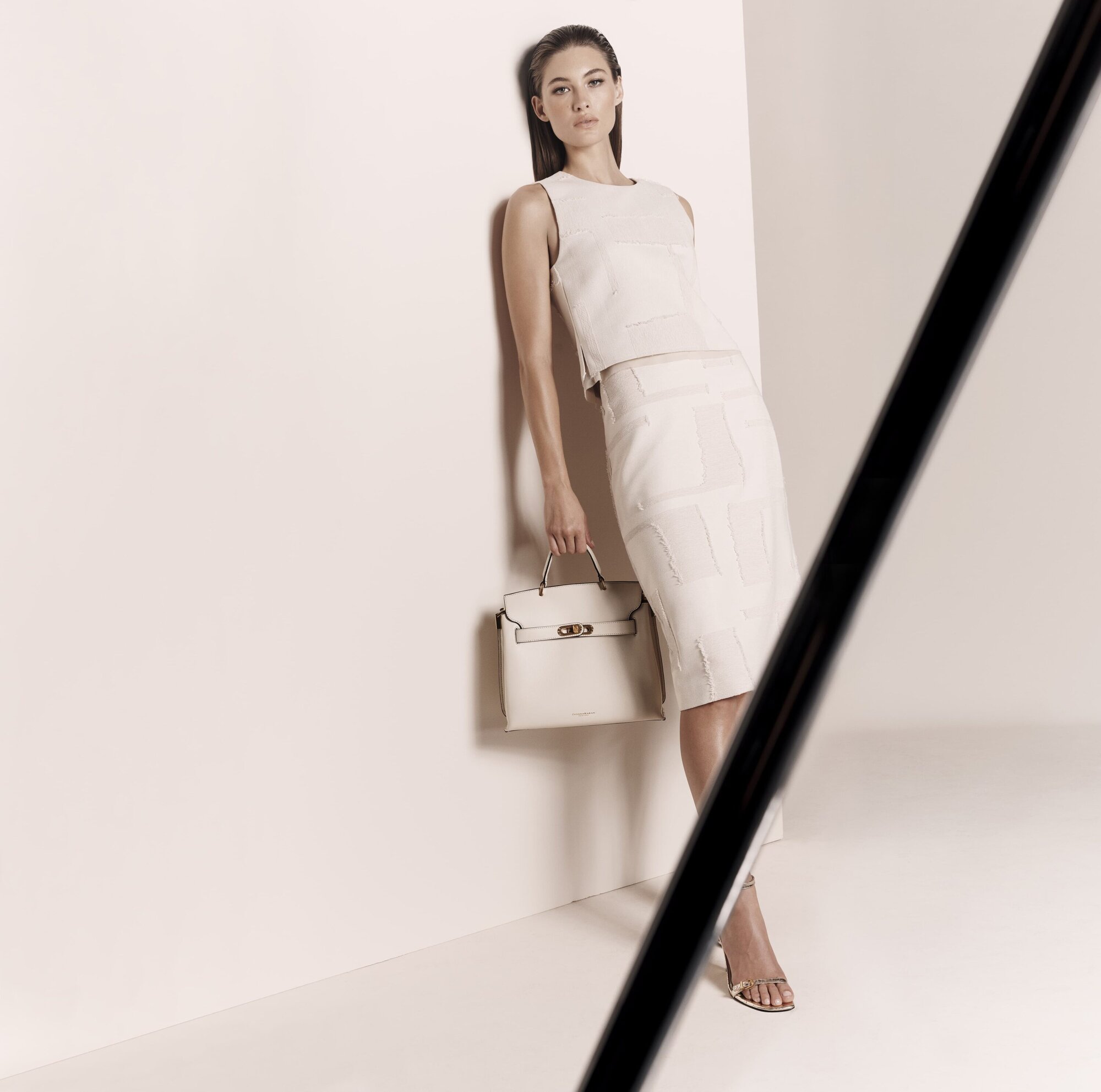 A poised woman in a cream-colored skirt suit holding a handbag stands against a light background, bathed in soft, studio lighting.