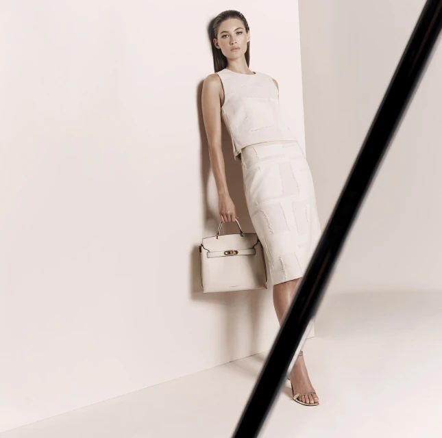 A poised woman in a cream-colored skirt suit holding a handbag stands against a light background, bathed in soft, studio lighting.