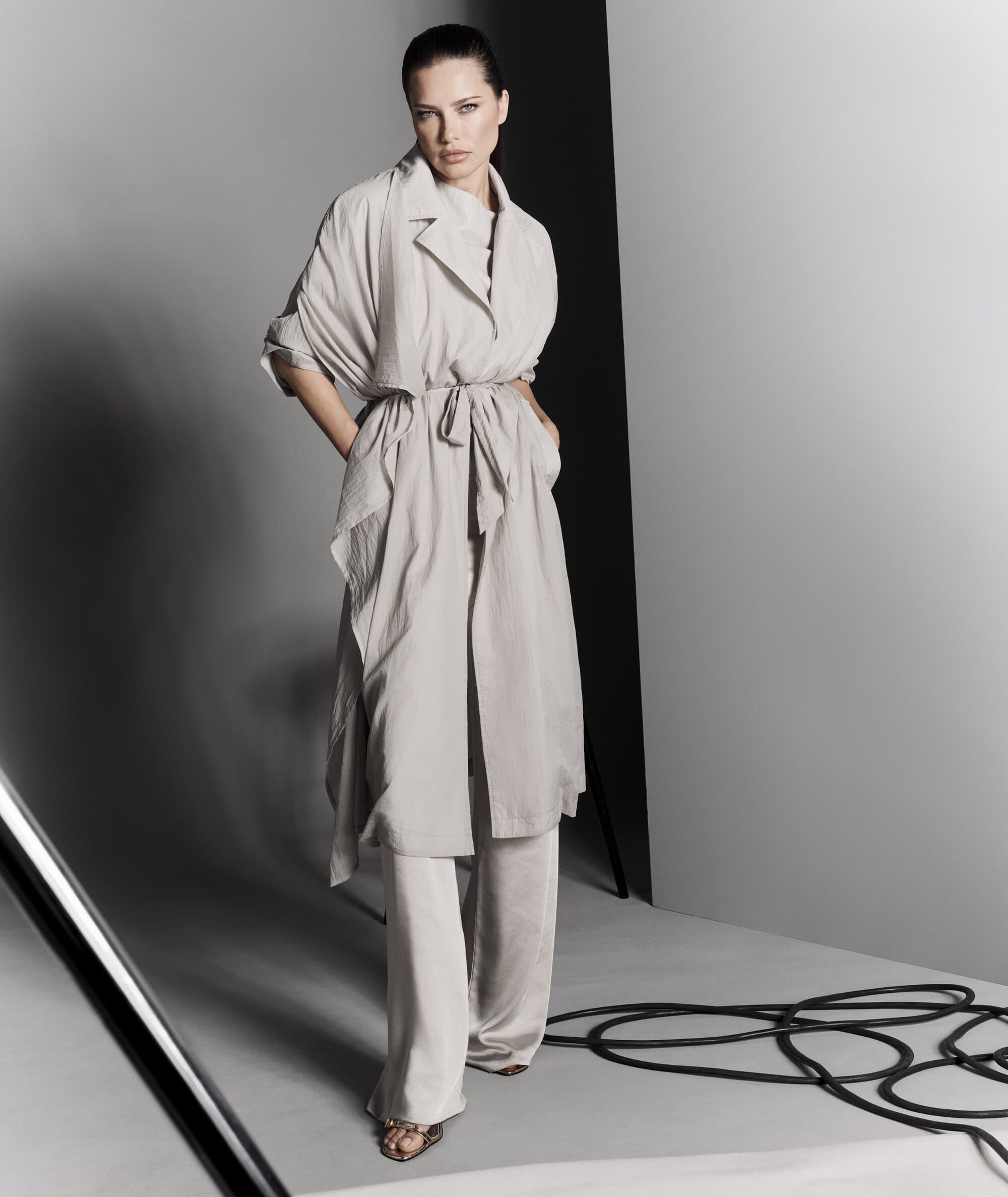 Full-length shot of a woman in a light-colored belted coat and pants posing in a studio with gray walls and black cables on the floor.